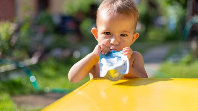 serious baby eating fruit puree in baby food pouch and looking into the camera in front of the yellow table. on the background is a green garden on a sunny day in blur