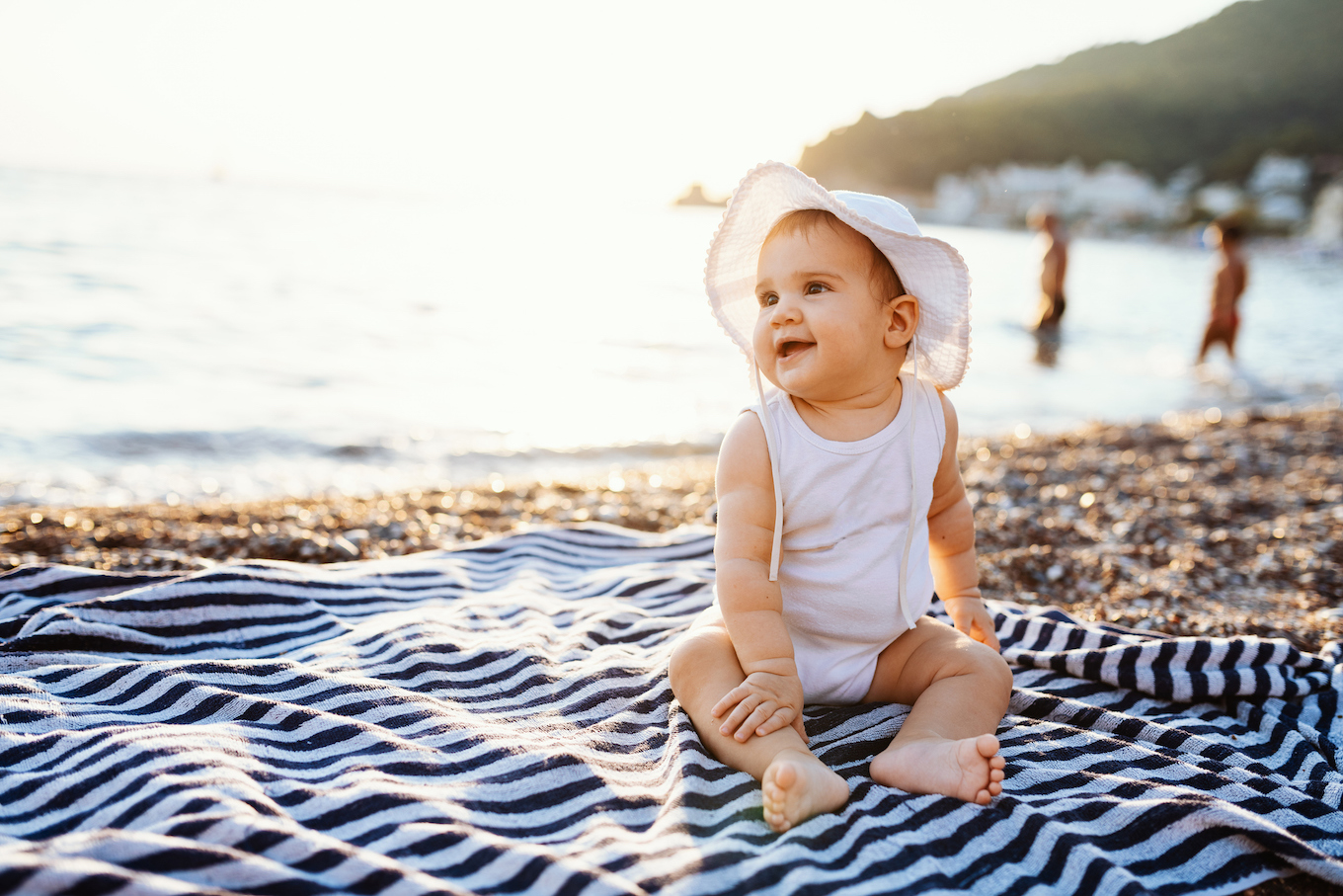 Baby girl with hat sitting on towel at the beach in summer
