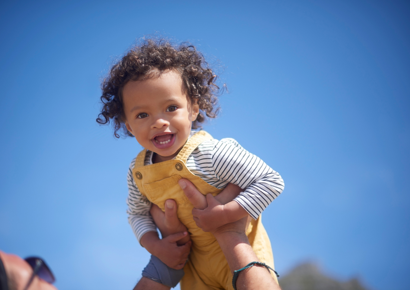 Happy, laughing and portrait of child with a parent on a blue sky, bonding and playing in nature. Smile, carefree and face of a little girl being held by a person with love, care and playful in summer