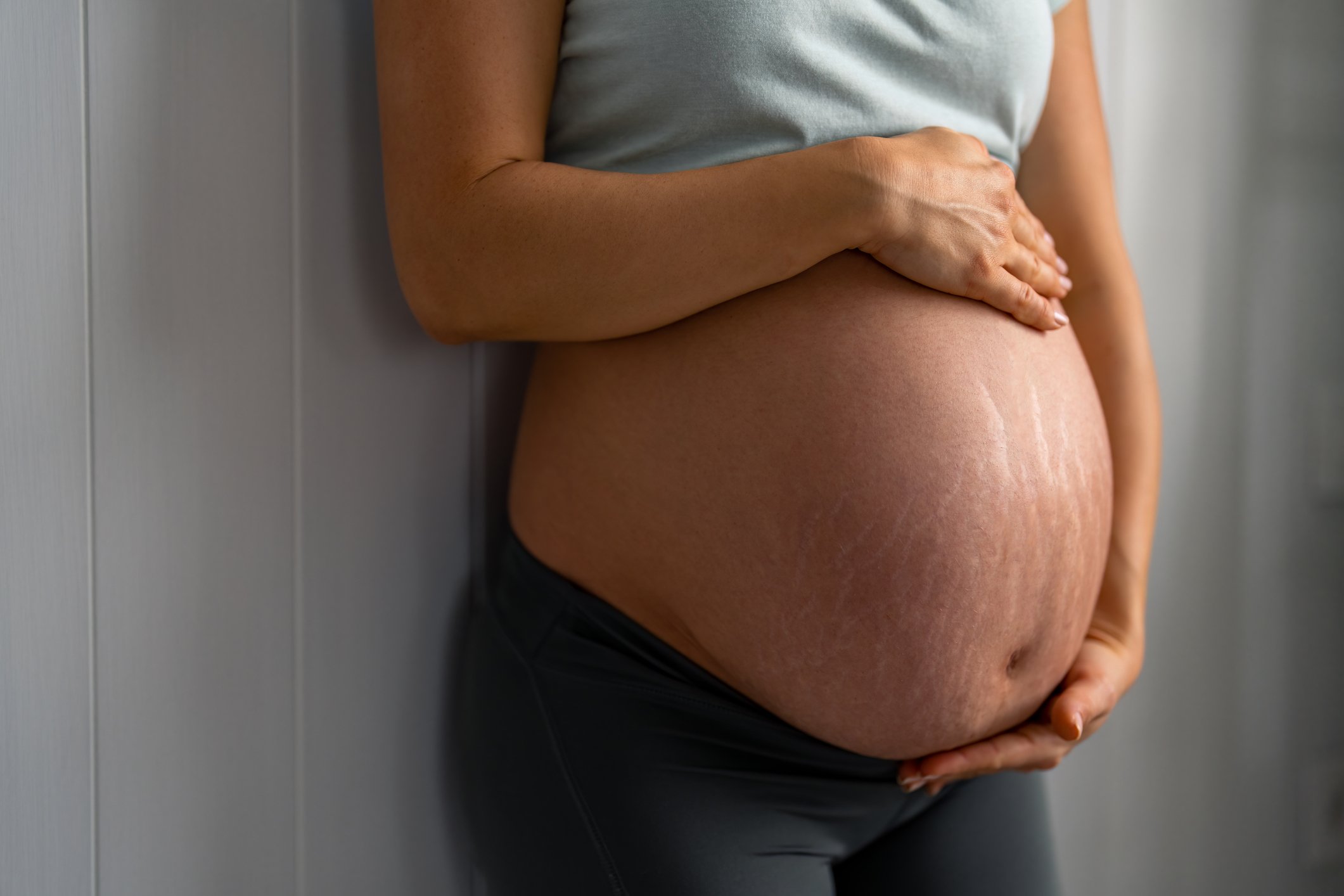 Unrecognizable pregnant woman with stretch marks wearing comfortable clothes, soft blue t-shirt and dark gray leggings, holding her hands on stomach, standing indoors against gray wall.