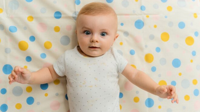 White baby wide-eyed laying on colorful sheet.