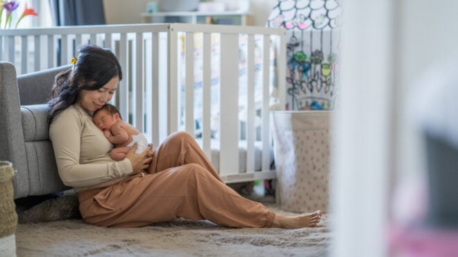 A new Mother sits on the floor of her daughters nursery as she takes a moment to enjoy Motherhood. She is dressed comfortably and holding her baby close as the two bond.