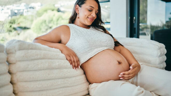 A joyful pregnant woman with long dark hair, sitting on a cozy sofa in a bright living room, gently cradling her belly and smiling warmly.